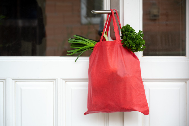 Das Bild zeigt einen roten Einkaufsbeutel an einer Klinke hängend, gefüllt mit Gemüse