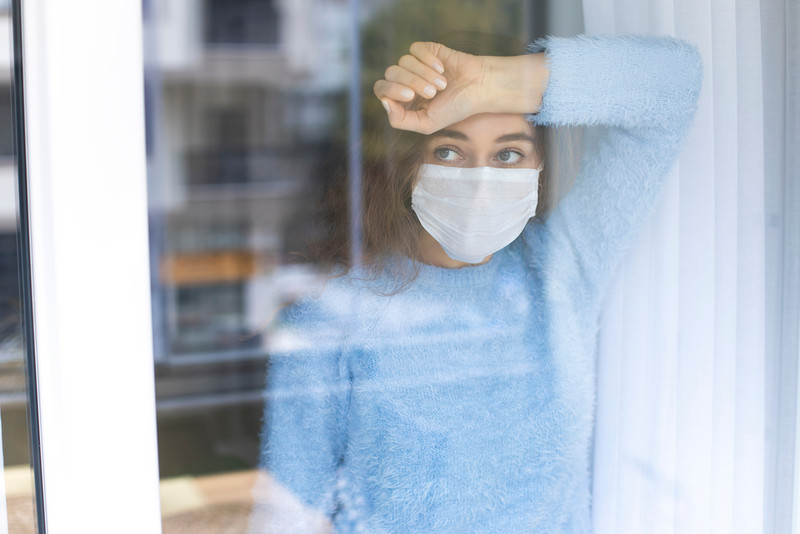 Das Bild zeigt eine Frau mit Maske hinter einem Fenster.
